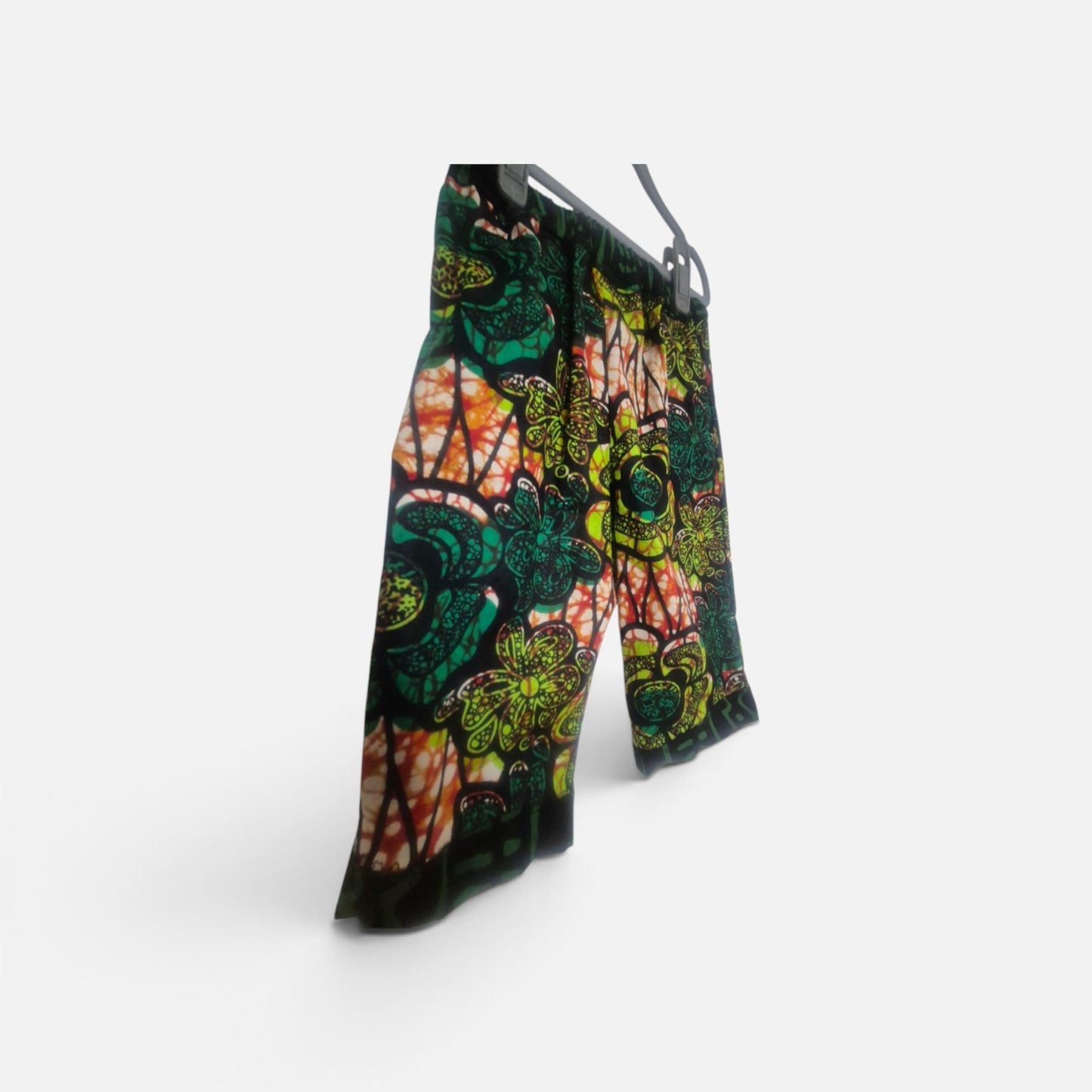 A pair of wide-leg children's trousers featuring a vibrant, multicolored batik-style floral and abstract pattern in green, orange, yellow, and black, hanging on a minimalist gray hanger against a clean white background.