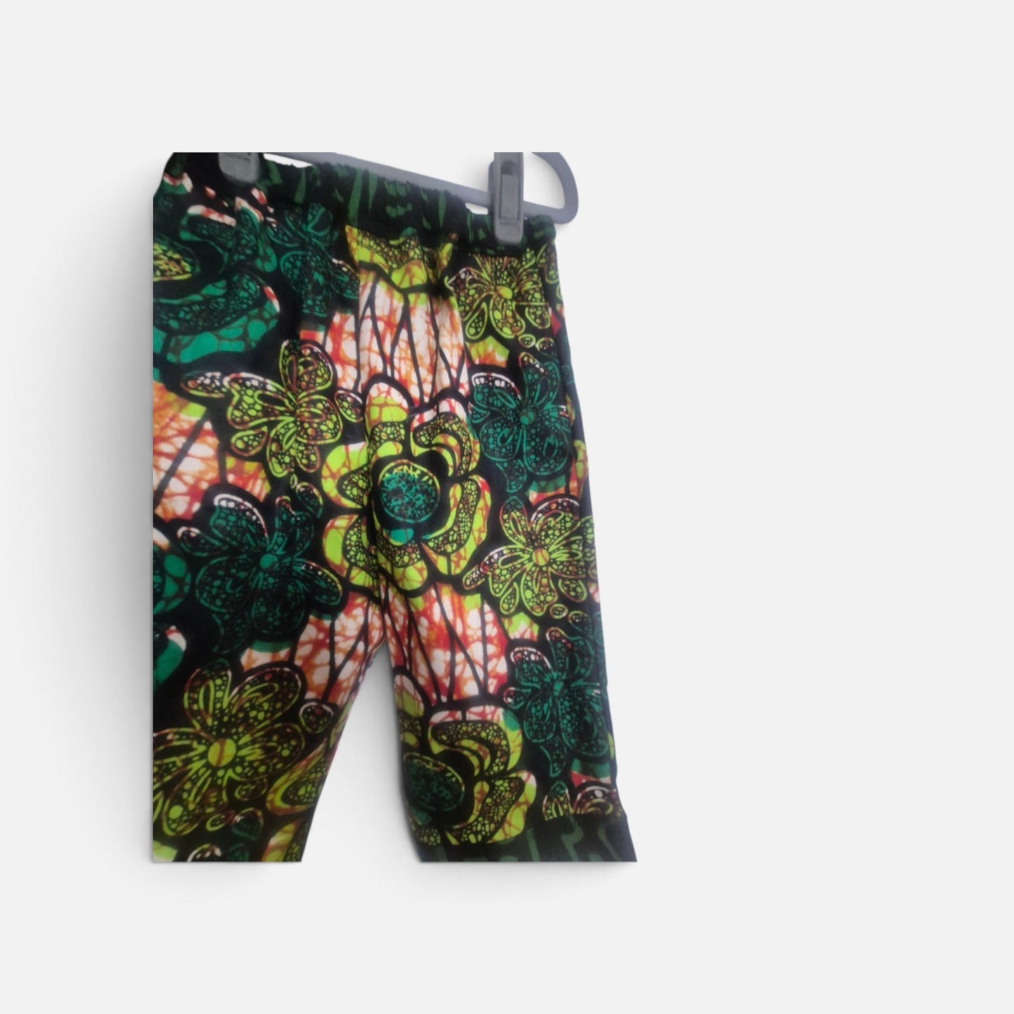 A pair of children's trousers featuring a vibrant, multi-color African wax print pattern with greens, white, red, and black,and floral motifs, hanging on a gray plastic flocked hanger against a light gray background.