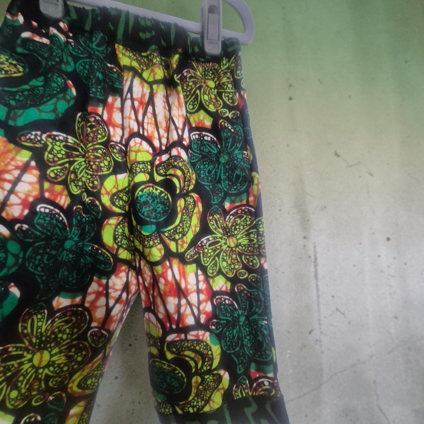 A pair of children's tousers with a vibrant, multi-colour batik-style floral pattern in dark and lime green, red,  white, and black, hanging on a grey plastic hanger against a green wall.