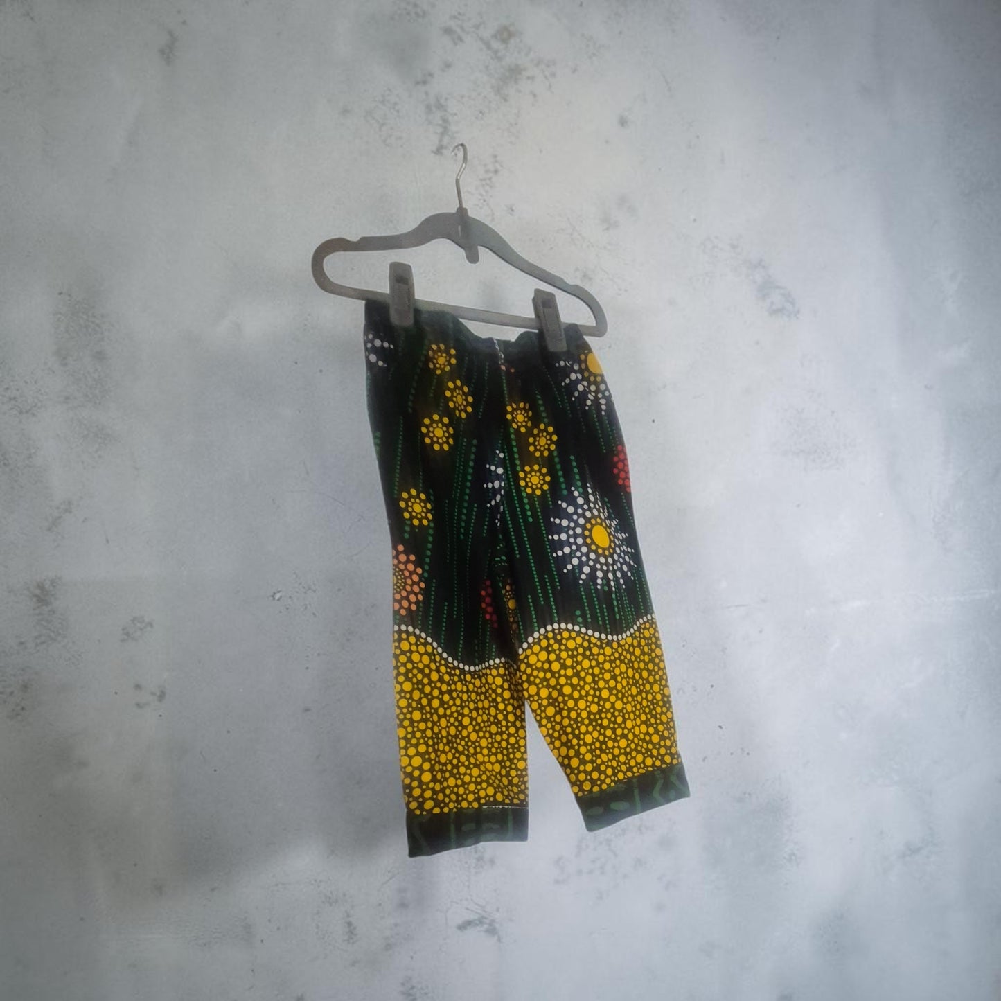Black cropped pants with a vibrant yellow, white, and red dot-art floral pattern, hanging on a gray hanger against a textured gray wall.