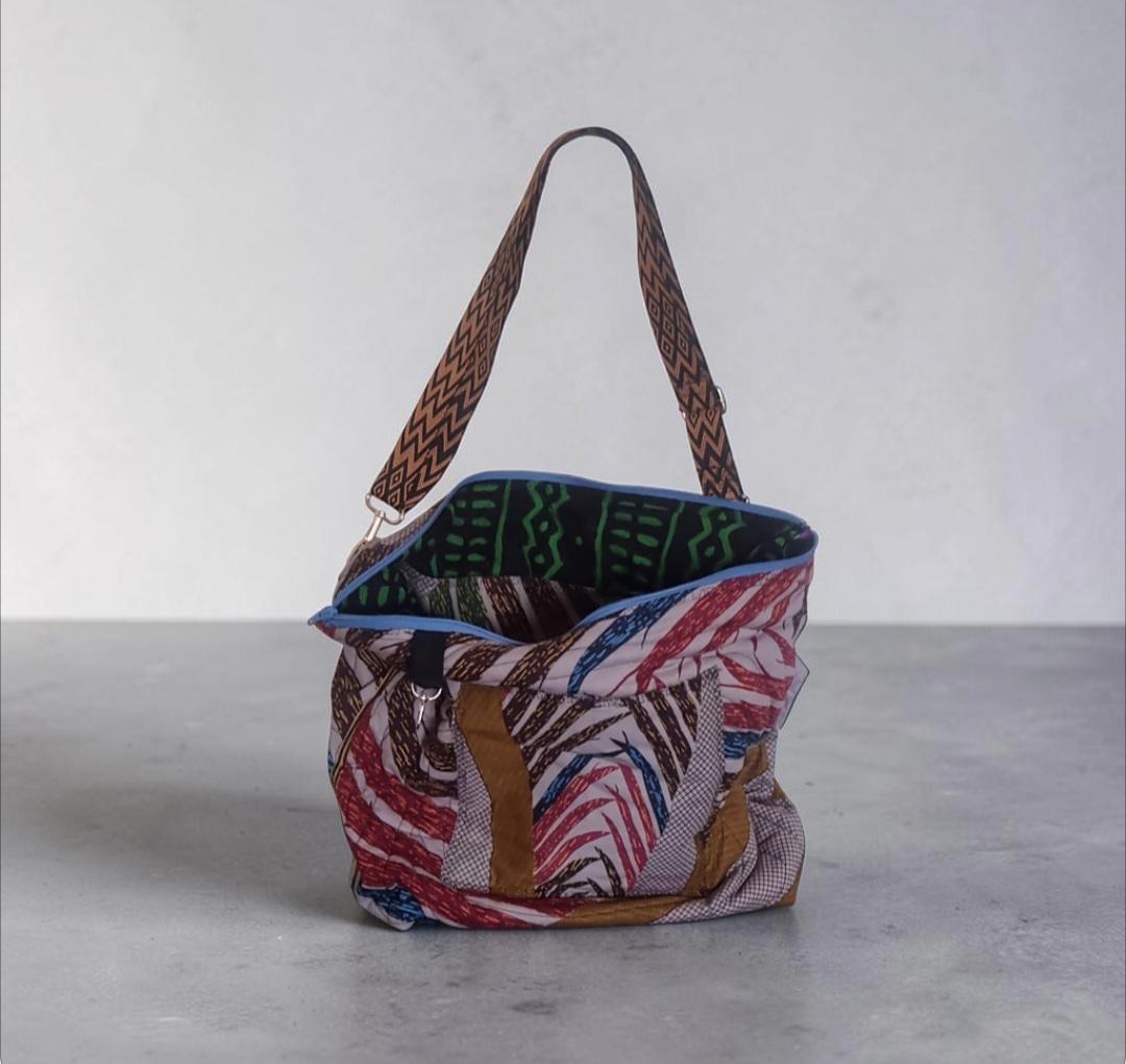 A handmade shoulder bag with a multicolor African wax print fabric body, brown patterned straps, and a green patterned lining, set against a neutral gray background.
