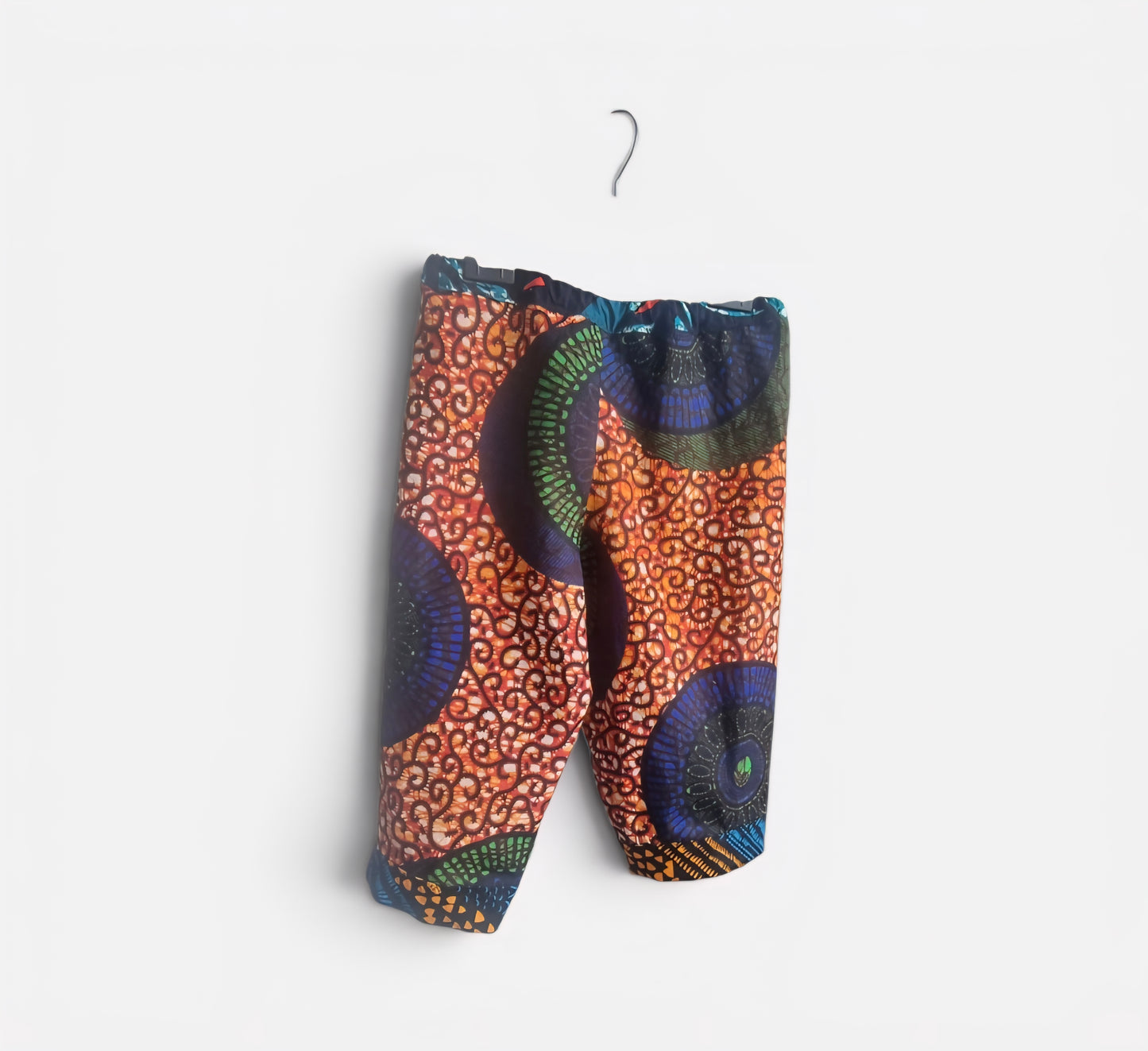 A pair of children's shorts with a vibrant African wax print pattern, featuring orange, blue, green, and black geometric and circular motifs, hanging on a small metal hanger against a plain white background.