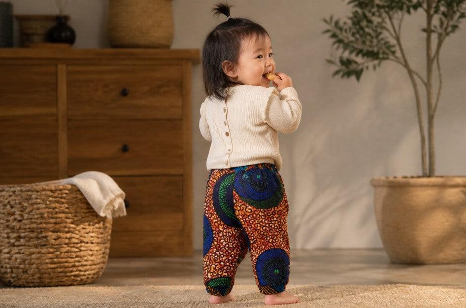 A toddler standing indoors wearing cream ribbed knit top and colorful patterned pants with blue, green, orange, and red circular motifs, photographed in a warm neutral room with a wooden dresser, basket, and potted plant.