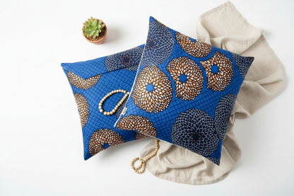 Flat lay of two blue and gold African wax print envelope cushion covers with a small plant and linen
