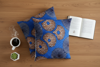 Flat lay of two blue and gold African wax print cushion covers on oak floor with coffee and book