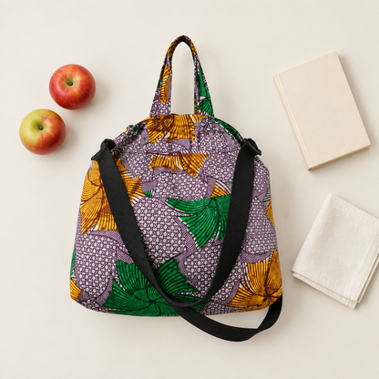 Fans and Dots African wax print market bag with black shoulder strap, arranged beside apples and a book on a pale background.