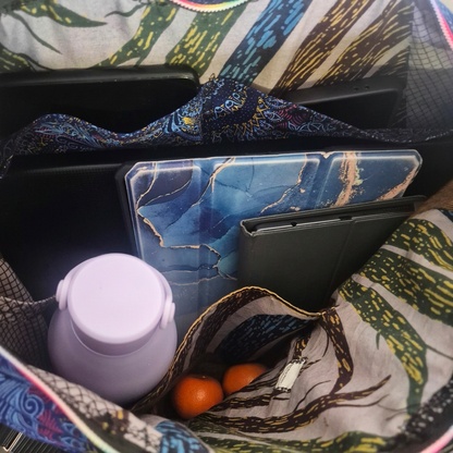 Inside the Blue Peacocks Traveller, showing leaf-print lining and patch pockets holding a large laptop, eReaders, iPad, phone, a lavender water bottle and small fruits.